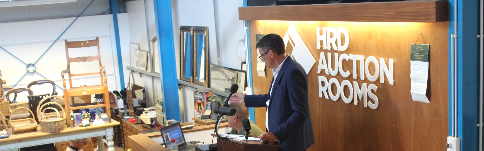 A person in a dark suit speaks at a wooden podium with microphones in front of the HRD AUCTION ROOMS sign, with chairs, baskets and frames on display nearby.