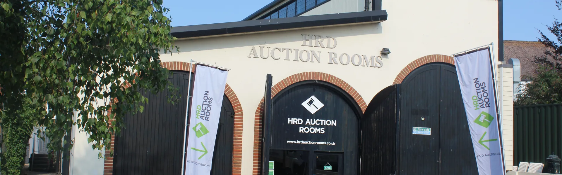 Cream building with brick-arched entrances and black doors, labelled HRD AUCTION ROOMS; two white banners stand nearby, a tree on the left and blue sky.