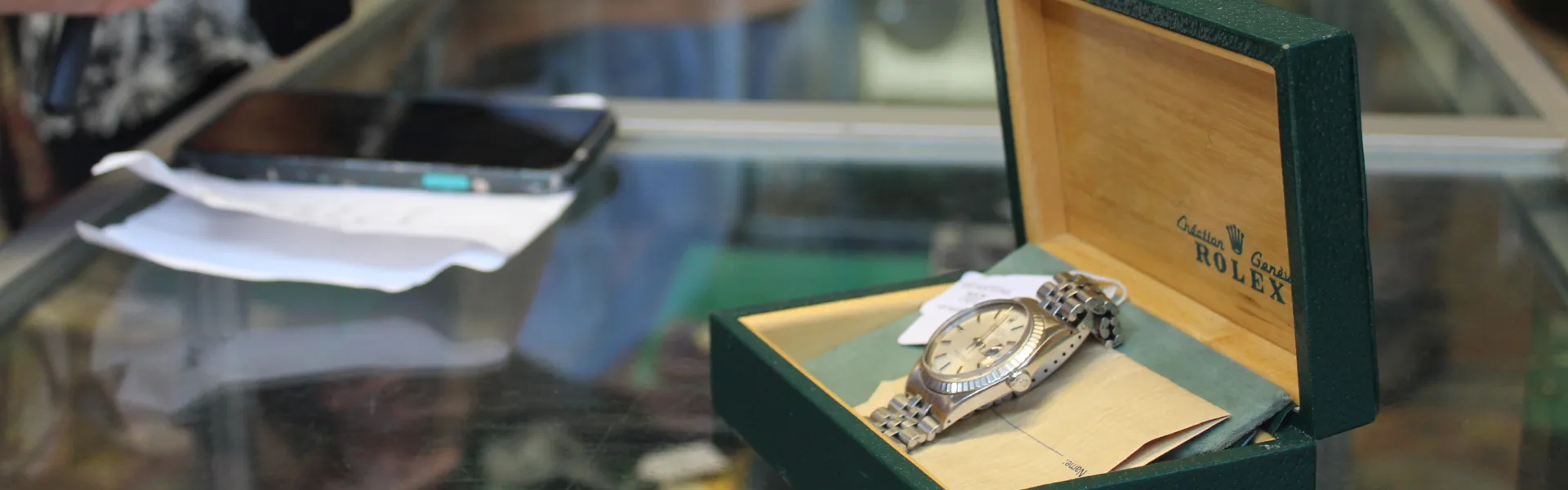 Open green Rolex box on a glass counter, displaying a silver Rolex with a cream dial; blurred background shows a smartphone and papers.