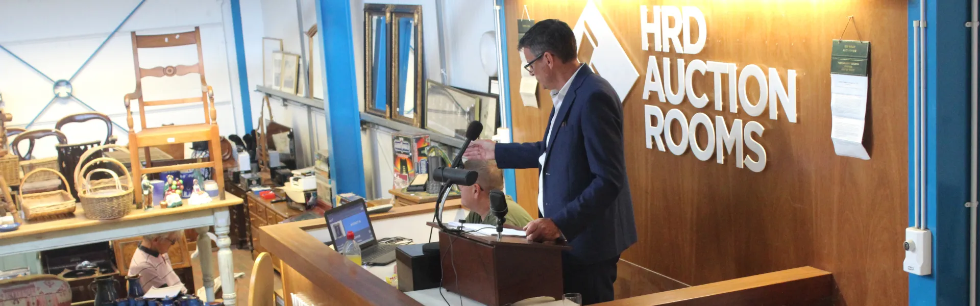 A person in a blazer speaks at a wooden auction podium with two microphones; HRD AUCTION ROOMS sign on a wood-panelled wall, surrounded by antiques.