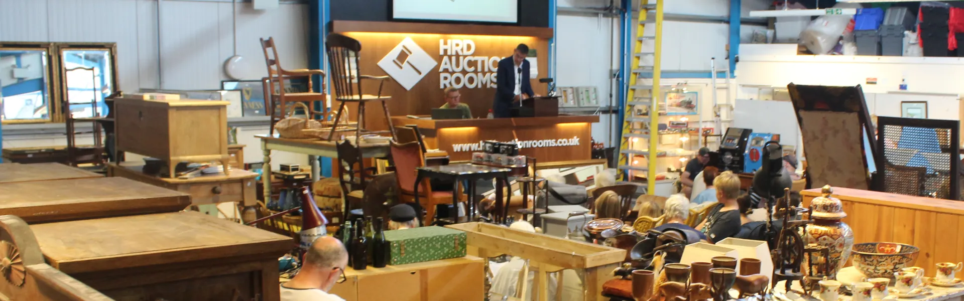 Interior of an auction hall crowded with antique furniture, pottery and decor on tables; a raised desk reads 'HRD Auction Rooms' as attendees browse.