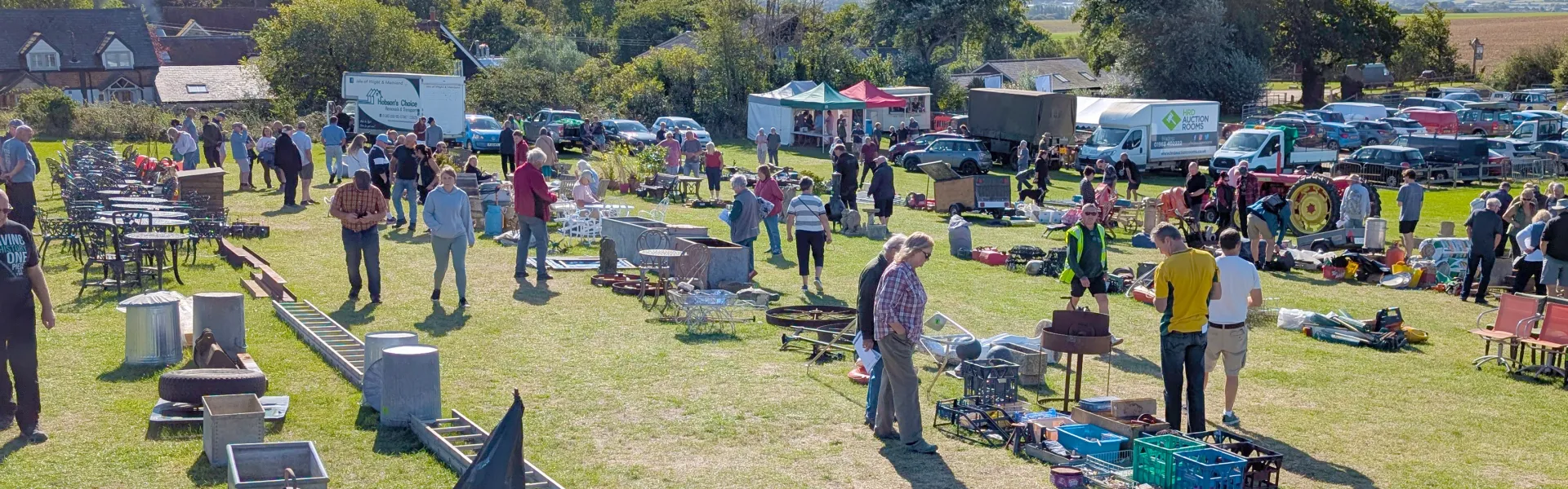 Outdoor flea market on a sunny day; people browse a grassy field of tables and items, with tents, stalls, and many parked cars in the background.