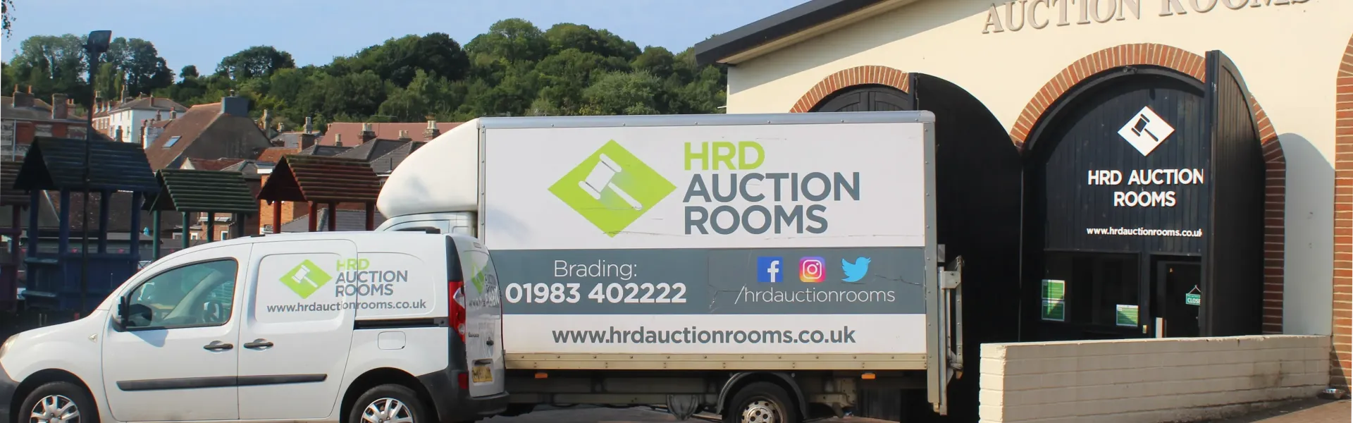 White HRD Auction Rooms van and a large branded trailer parked outside a beige auction building with brick arches and dark arched doors.