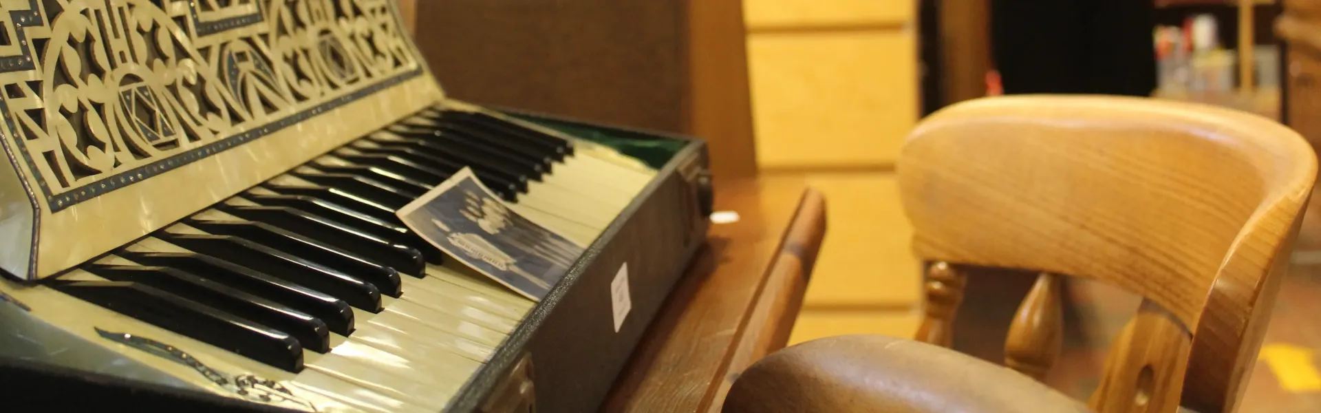 Close-up of an ornate keyboard instrument with black and white keys, decorative metal grille, a brochure lying on the keys, and a wooden chair beside it.