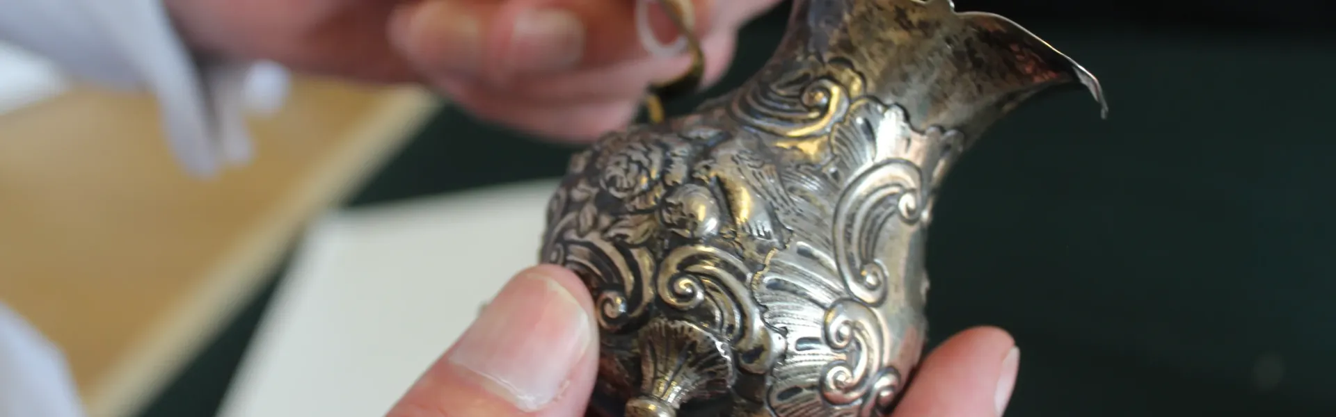 Close-up of a person holding an intricately engraved silver vessel with floral scrollwork and a curved spout.