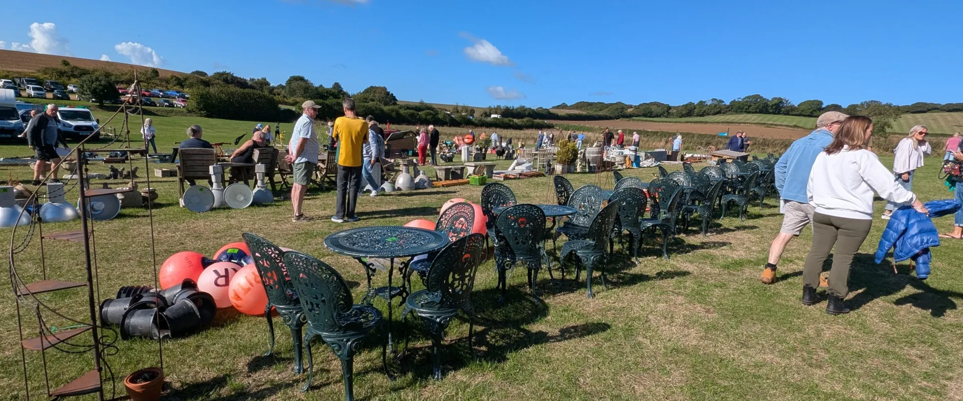 Row of decorative cast-iron chairs with matching tables on a grassy field at a sunny outdoor market, with stalls and shoppers in the background.