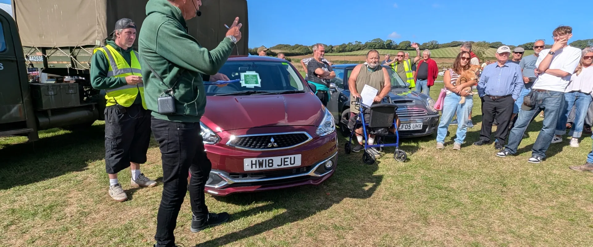 Outdoor car show with a person speaking into a headset mic beside a maroon car (HW18 JEU); crowd in hi-vis vests and casual clothing on a grassy field under a blue sky.