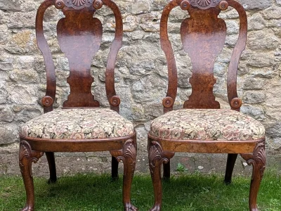Pair of antique carved wooden chairs with floral upholstered seats, featuring shell motifs on the backs and claw-foot legs, set on grass against a stone wall.