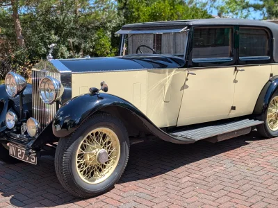Vintage luxury car with cream and black two-tone body, chrome grille and large round headlights, cream wire wheels, parked on a brick driveway.