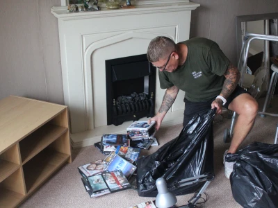 Person with tattoos in a green T-shirt and glasses sorting a pile of DVDs into a black trash bag in a living room by a fireplace.