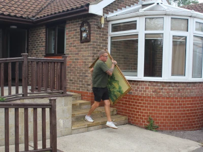 Person in a green shirt and black shorts carries a large framed landscape painting down stone steps beside a brick house with a white bay window.