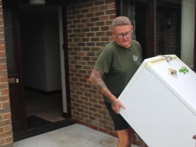 Person with short hair, glasses, and tattoos in a green shirt carries a white refrigerator/freezer out of a brick house.