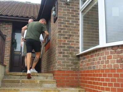 Two people with visible tattoos carry a large white appliance up concrete steps to a brick house entrance; bay window and brick walls frame the scene.