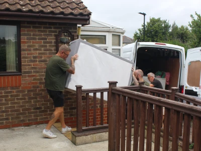 Three people unload a mattress from a white van outside a brick house; one person in a green shirt carries it while two others help nearby.
