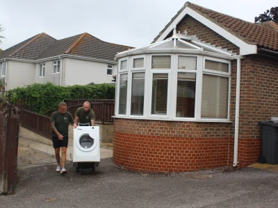 Two people in green shirts push a front-loading washing machine on a dolly along a gravel driveway beside a brick house with a white bay window.