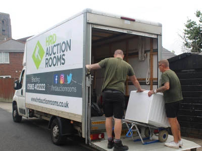 Two people load a white appliance into an HRD Auction Rooms van on a residential street; the van displays branding and contact details.