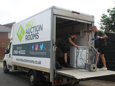 Two people unload a large appliance from a branded HRD Auction Rooms truck onto a ramp in a residential street.