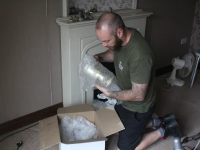 Bearded person kneels on a carpet, unwrapping a bubble-wrapped item from a cardboard box in a living room with a white fireplace.