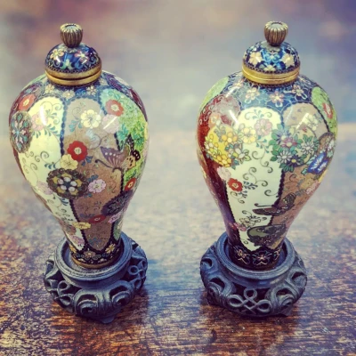 Two ornate cloisonné-style vases with lids on carved dark bases, decorated with vibrant floral patterns; placed on a wooden surface.