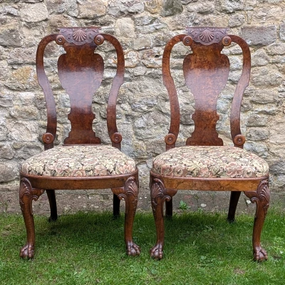 Pair of antique carved wooden chairs with floral upholstered seats, featuring shell motifs on the backs and claw-foot legs, set on grass against a stone wall.