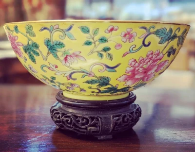 Yellow porcelain bowl with pink and teal flowers and scrolling vines on a dark carved stand, resting on a wooden surface.