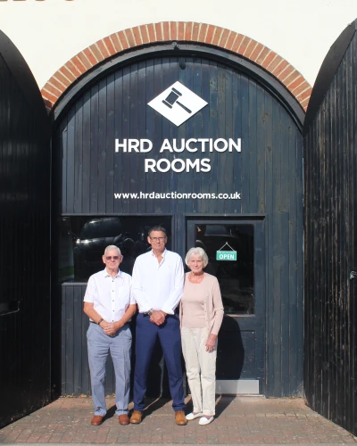 Three people stand outside HRD Auction Rooms in front of a dark wooden arched doorway with brick surround; an OPEN sign on the door.