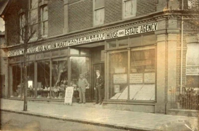 Sepia street view of a brick building with large shopfronts, a sign reading House and Estate Agency; two people stand on the pavement beside a board.