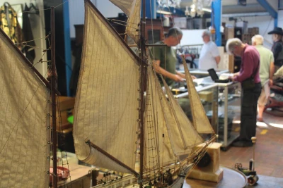 Close-up of a model sailing ship with beige sails and rigging; blurred background shows several people at a workshop counter.