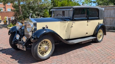 Vintage luxury car with cream and black two-tone body, chrome grille and large round headlights, cream wire wheels, parked on a brick driveway.