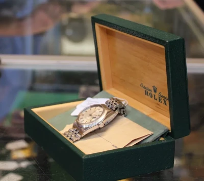 A silver Rolex watch with a metal bracelet sits in an open green presentation box with a beige interior and Rolex branding.
