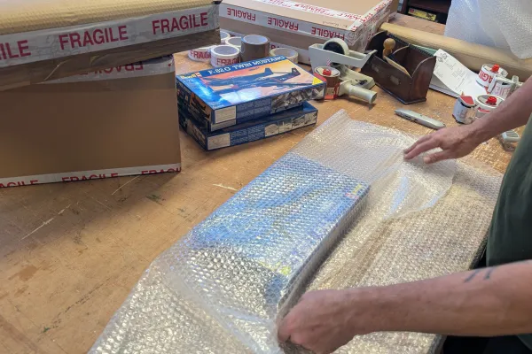 A person in a green shirt wraps a long item in bubble wrap on a cluttered workbench, with fragile boxes, tape rolls and model kit boxes nearby.