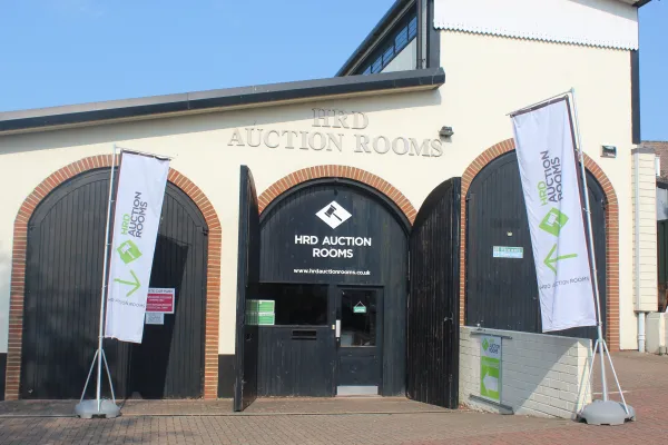 Exterior of HRD Auction Rooms: cream building with brick-arched doorways, central open black doorway, flanked by tall banners; blue sky above.