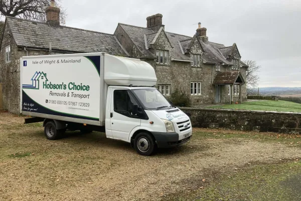 White Hobson's Choice removal truck with Isle of Wight branding parked on gravel beside a stone farmhouse; cloudy sky overhead.