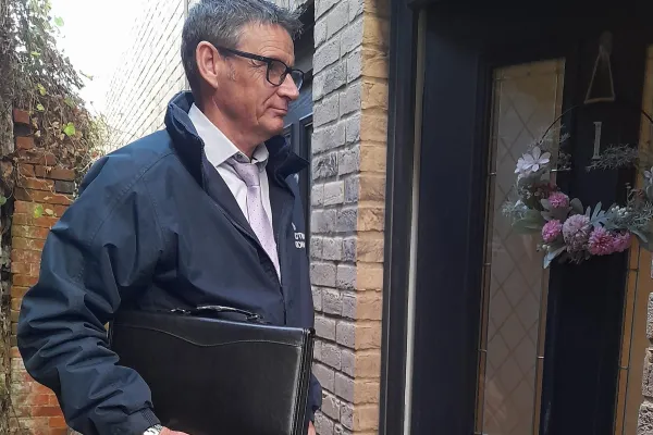 Person in a navy jacket, white shirt and pink tie, wearing glasses, holds a black folder beside a brick doorway with a floral wreath.