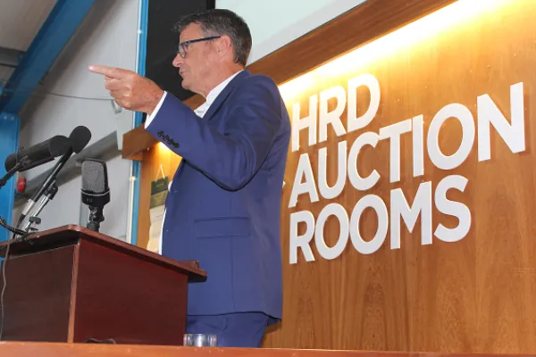 Person in a blue suit at a wooden podium with microphones, gesturing with a pointed finger, in front of a wall with large "AUCTION ROOMS" lettering.