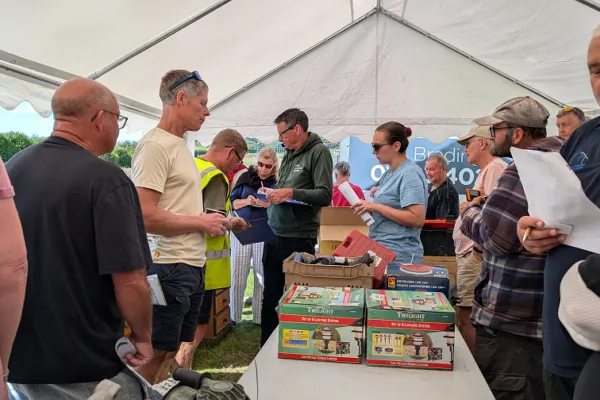 Group of diverse people under a white canopy gathers around a long table with boxed items and papers, exchanging goods at an outdoor event.