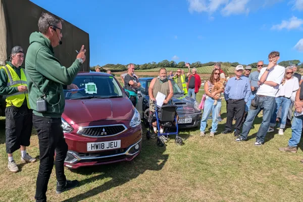 Speaker in a green hoodie with a headset mic addresses a crowd beside a red car on a sunny grassy field.