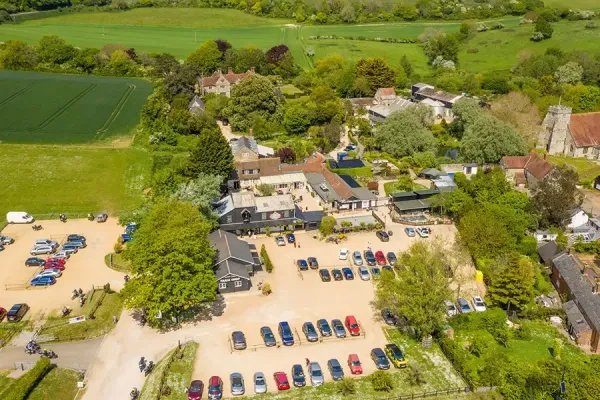 Aerial view of a rural village with buildings around a central square, surrounded by green fields and trees; a large car park is filled with colourful cars.