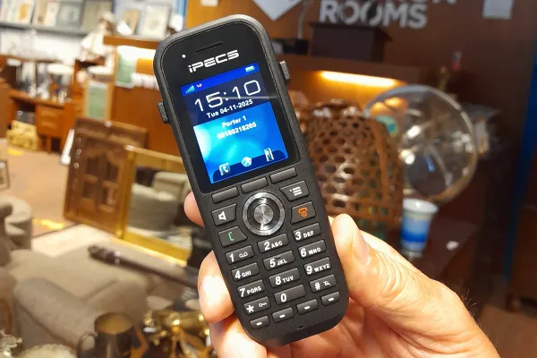 Hand holding a black iPECS mobile phone with a blue screen displaying 15:10, in an auction room with wooden furniture and various displayed items.