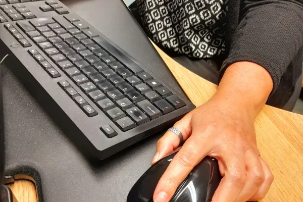 Close-up of a person typing on a black keyboard and using a black mouse on a wooden desk with a large mouse mat; wearing a black-and-white patterned top.
