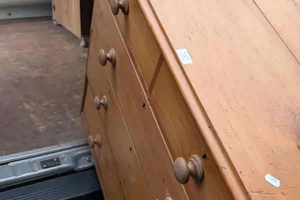 A wooden dresser with multiple drawers and round knobs is angled into a moving truck, with a hand gripping the bottom edge on the right.