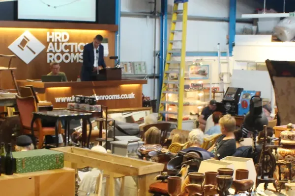 Warehouse auction: a person in a suit speaks at a raised podium while another sits nearby; HRD Auction Rooms sign visible as shoppers browse items.