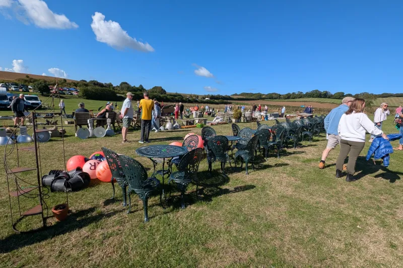 Row of decorative cast-iron chairs with matching tables on a grassy field at a sunny outdoor market, with stalls and shoppers in the background.