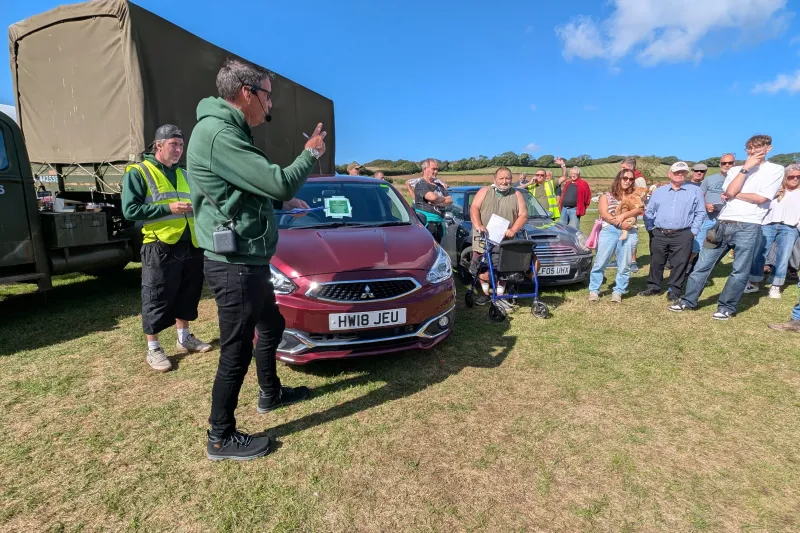 Outdoor car show with a person speaking into a headset mic beside a maroon car (HW18 JEU); crowd in hi-vis vests and casual clothing on a grassy field under a blue sky.